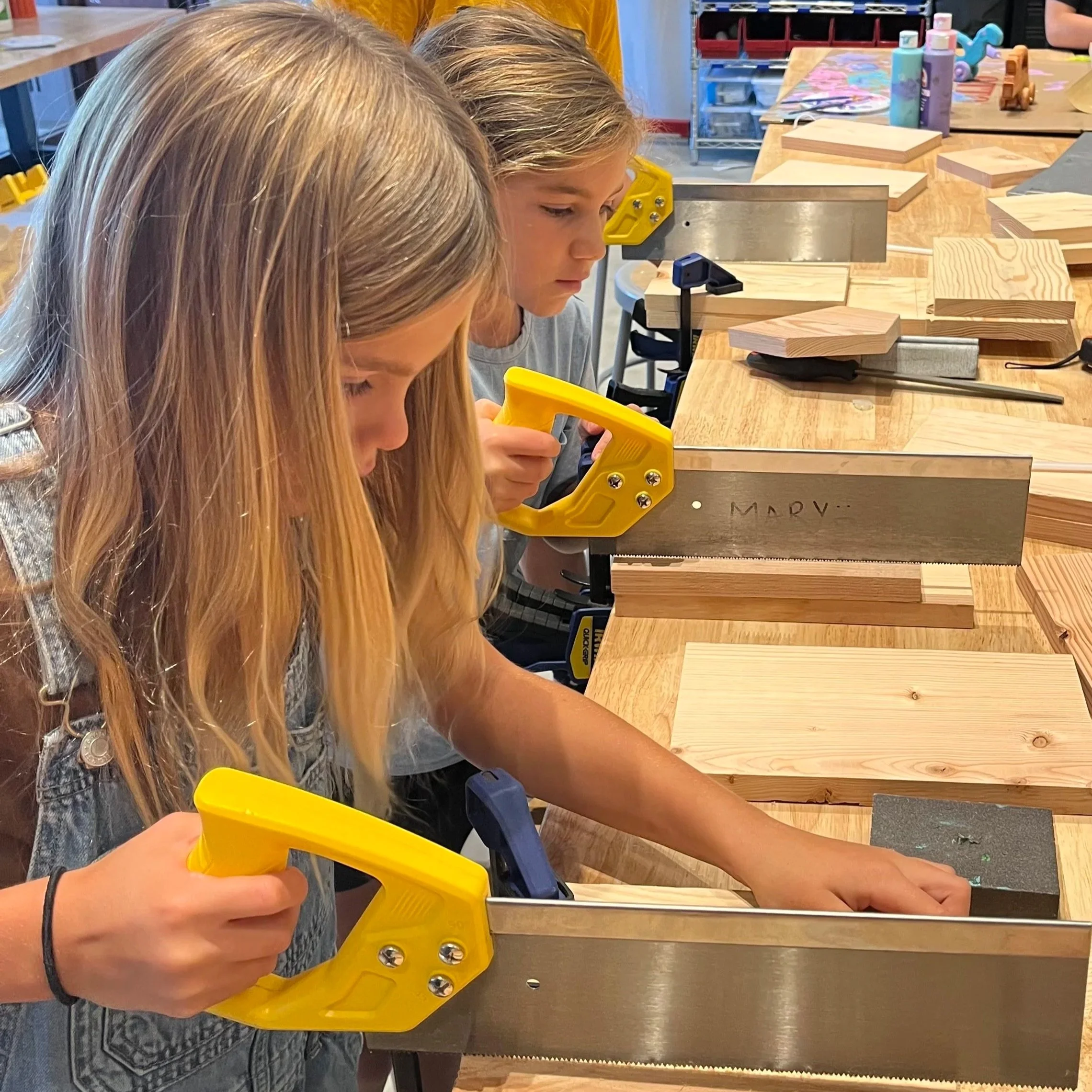 A kid using a clamp and saw to cut wood at a Forge day camp.